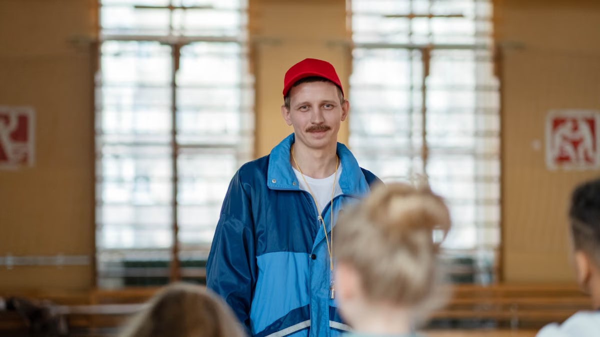 Male coach in a gym engaging with a group of children, wearing casual sportswear.